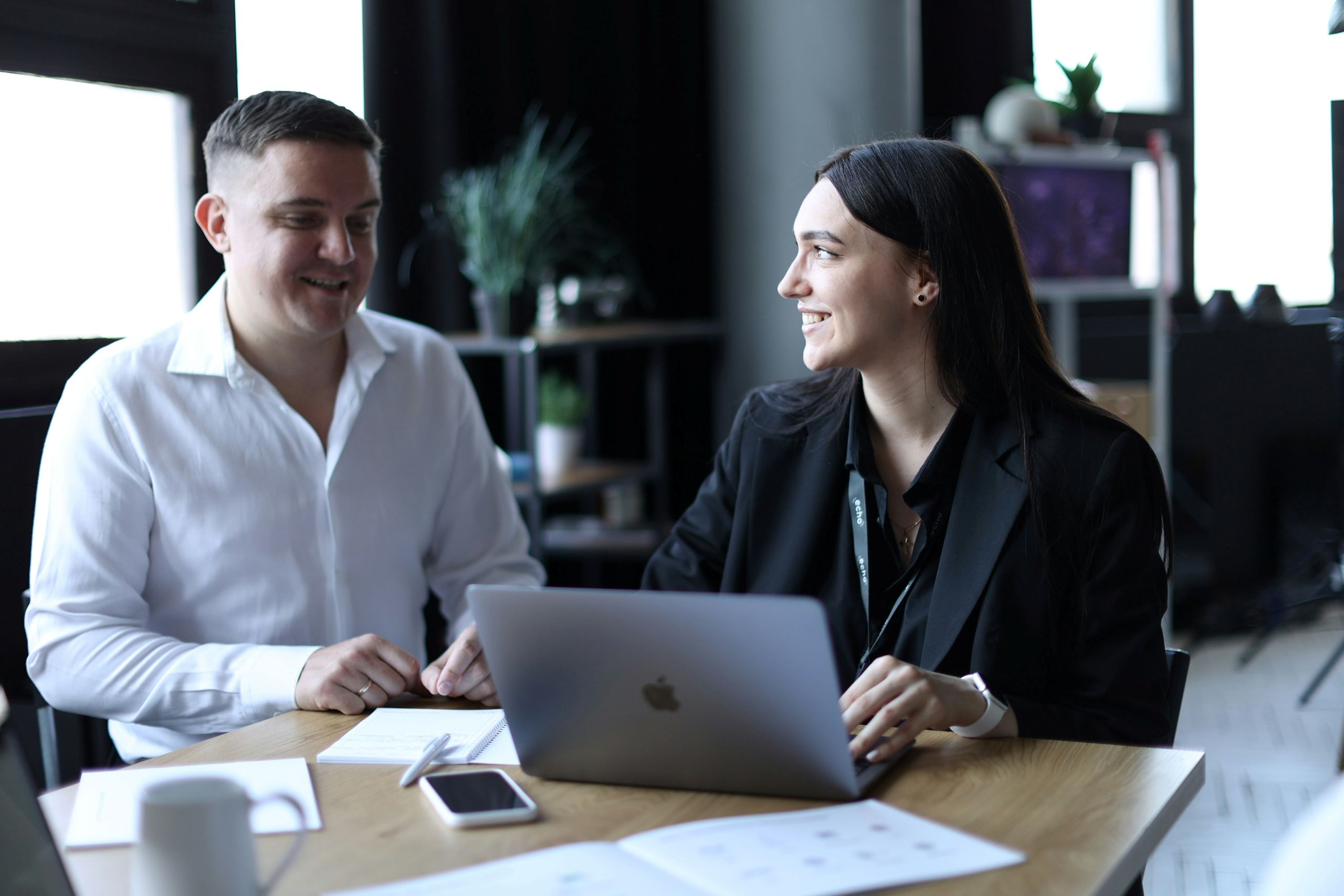 Two people smiling in front of desk with laptop and notebooks.
